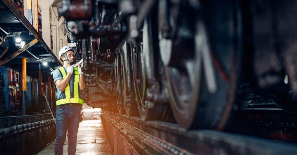 A railcar maintenance worker in a yellow vest inspects a railcar undercarriage.