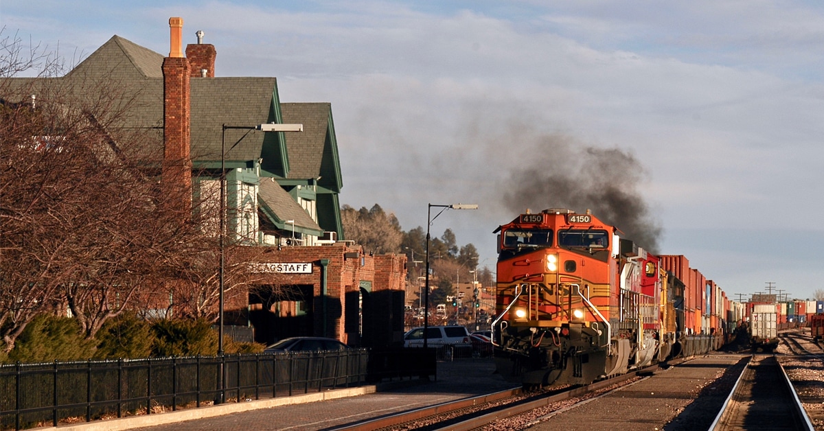 An orange and yellow rail freight train hauls rail cargo through a small town.