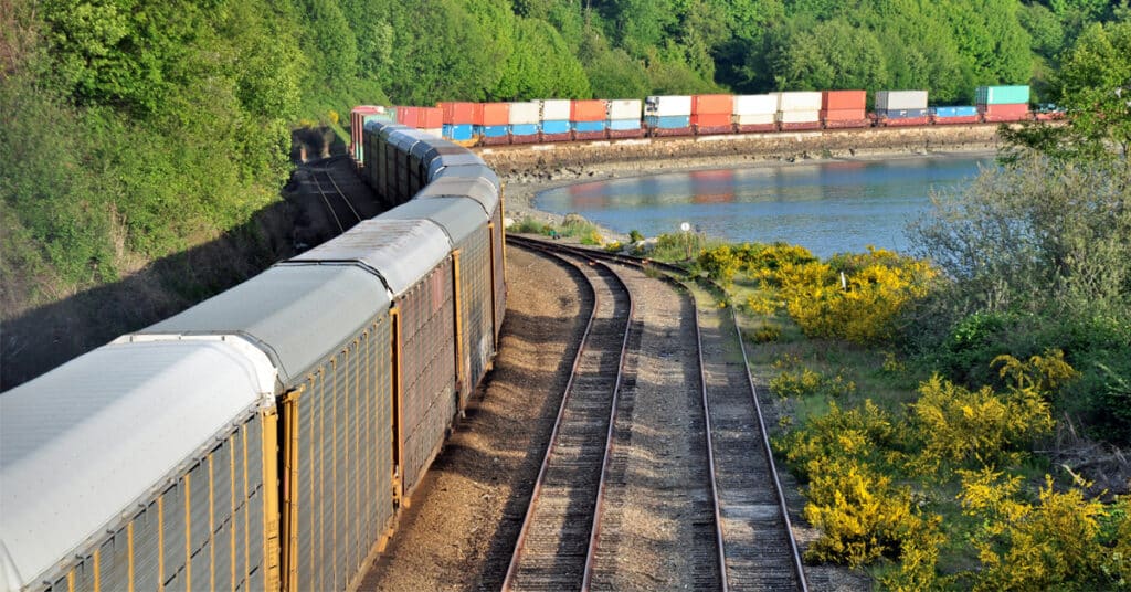 An autorack and freight train winds along rails along a river and forest.