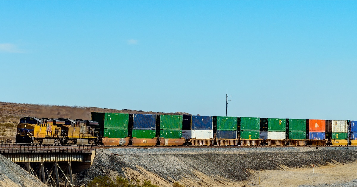 A yellow train hauls green and blue railcars through a shrub desert.
