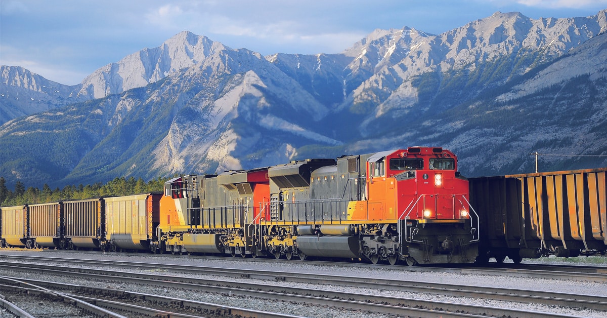A red, yellow, and black train hauling yellow cars on a track with snow covered mountains in the background.