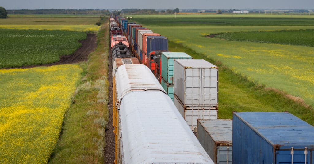 Two lines of rail shipping freight cars go through the middle of a green prairie and farmlands.