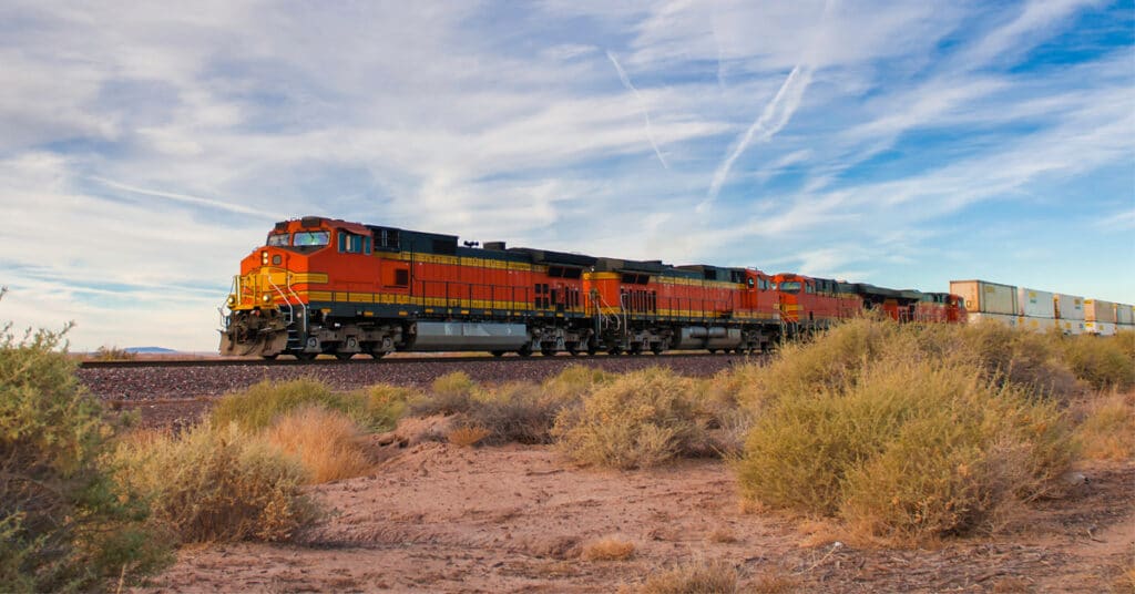 An orange rail freight engine hauls a line of rail shipping cars through a shrub desert.