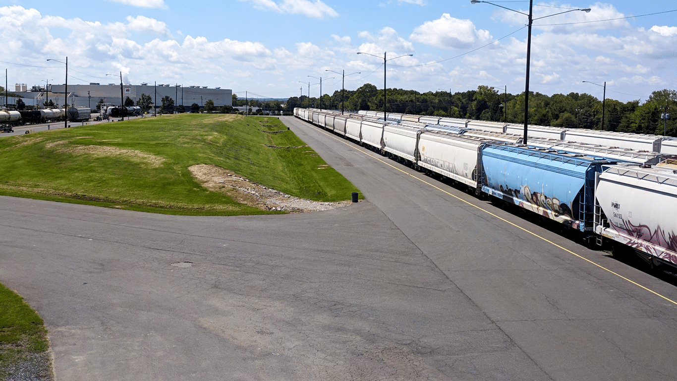 A series of blue and white railcars lined up on pavement next to some green grass and a factory.