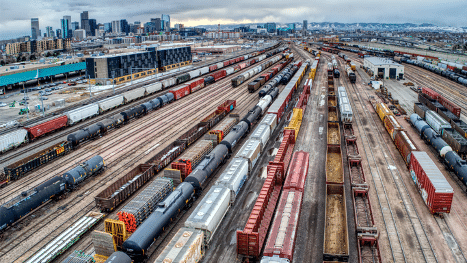 Aerial view of train switching yard with various railcars and, in the distance is the Denver Colorado skyline.