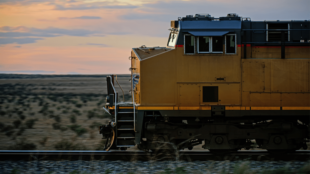 A yellow freight engine speeds across a shrub desert against a sunset.