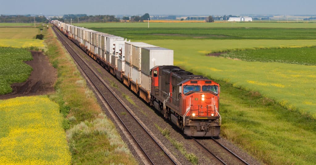 An orange rail freight engine pulls a train of rail cars through green farms.