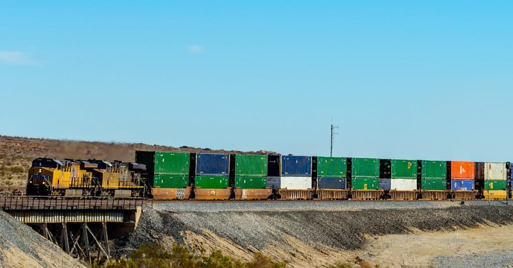 A yellow rail engine hauls a line of rail freight cars through a scrub desert.
