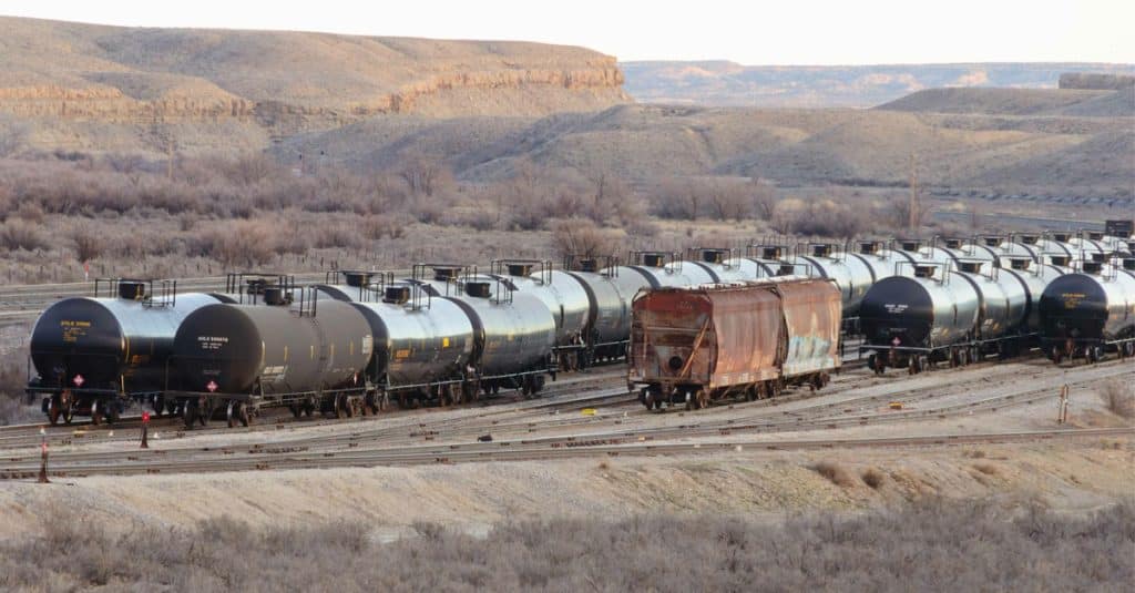 A yard of black rail freight oil tankers amidst shrubs and canyons.
