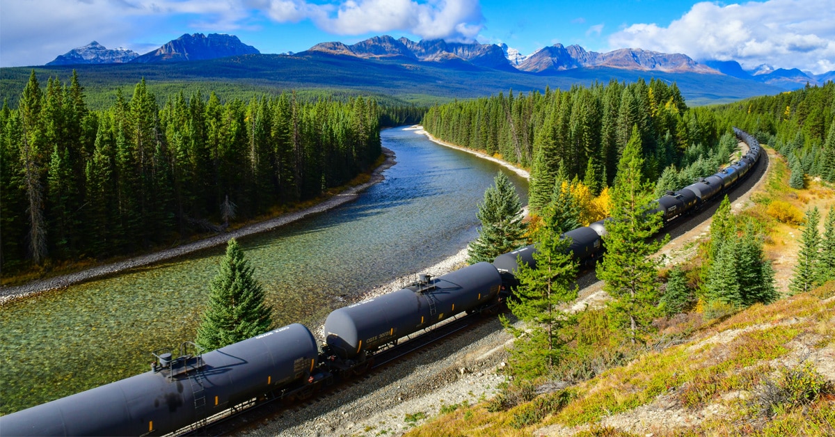 A train of rail tank cars moving through a forest along a river with mountains in the background.