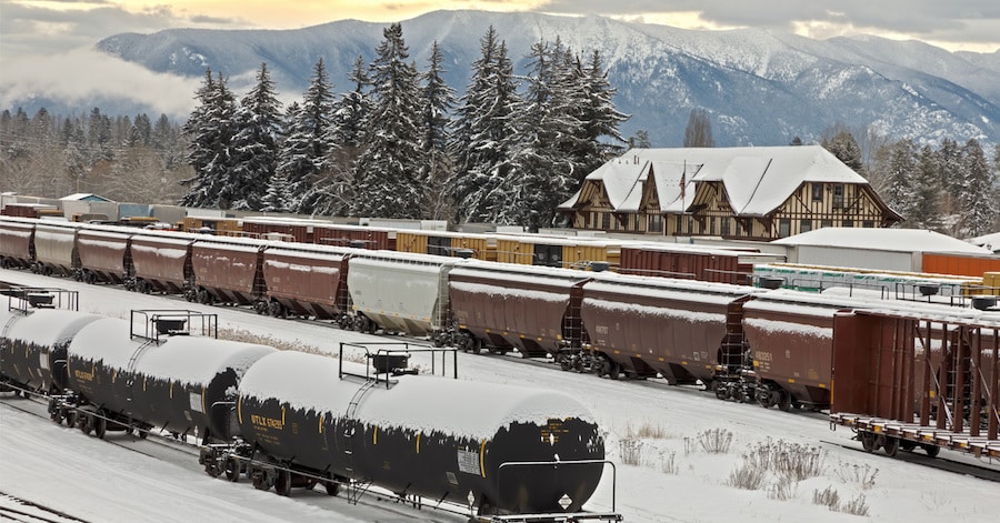 Rail yard in winter with snow covered trains, dept, and mountains in background.